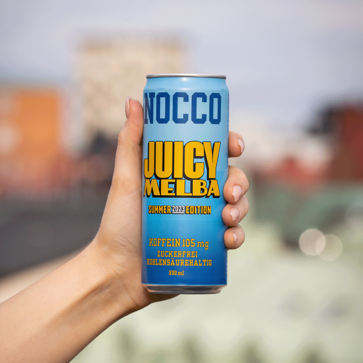 Hand hält eine Dose NOCCO "Juicy Melba" Summer 2023 Edition vor unscharfem Hintergrund.