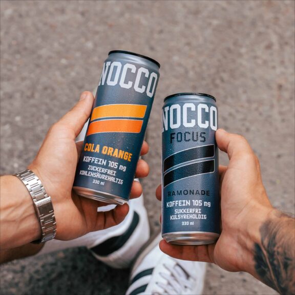 Zwei Hände halten NOCCO Energy-Drinks in den Geschmacksrichtungen Cola Orange und Ramonade.